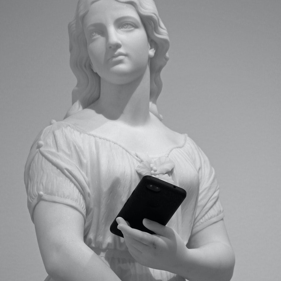 Low angle of stone classic statue with perfect skin and wavy hair holding modern cellphone near wall